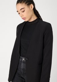 Svart blazer med skreddersydd design, enkelt knappelukking og en glatt tekstur. Båret over en svart høyhalset topp og kombinert med mørke jeans.