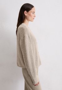 Beżowy, puszysty sweter z dzianiny o luźnym kroju, okrągłym dekolcie i opadających ramionach, zestawiony z jasnymi spodniami.