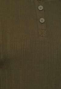 Olivgrünes, geripptes Oberteil mit dreieckiger Knopfleiste, das aus strukturiertem Stoff besteht und ein genähtes X-Detail in der Nähe der Knöpfe aufweist.