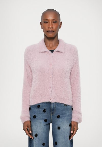 Cardigan en peluche rose clair avec un col et une fermeture à boutons, présentant une coupe cropped. Associé à un jean bleu orné d'appliqués floraux noirs.