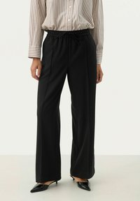 Pantalons larges noirs avec une texture douce, dotés d'une taille élastique et d'un cordon de serrage. Associé à une chemise à manches longues à rayures verticales.