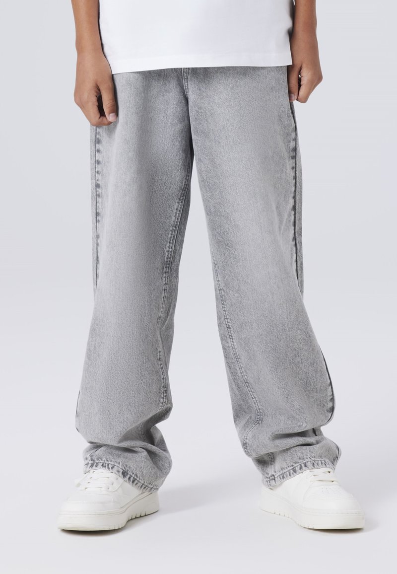 Svijetlosivi oversized traper hlače s istrošenom teksturom, nošene s bijelim tenisicama. Dizajn ima ravne nogavice i elastične manžete.