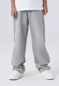 Svetlo sivi oversized jeans s poškodovano teksturo, obuti z belimi supergami. Oblikovanje ima ravne hlačnice in elastične manšete.