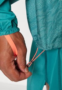 Cyan sportjack met een wit stippenpatroon. Bevat een verstelbare oranje trekkoord en elastische mouwen. Hand die het trekkoord aanpast.