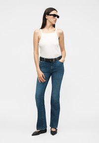 Γυναίκα με λευκό αμάνικο τοπ, μπλε τζιν bootcut με μαύρη ζώνη, μαύρα μυτερά παπούτσια και μαύρα γυαλιά ηλίου, στέκεται έχοντας το ένα χέρι στην τσέπη.