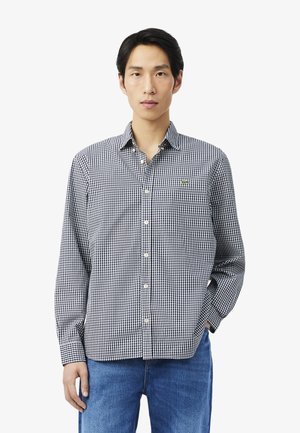 Joven con camisa de cuadros en blanco y negro y pantalones vaqueros azules, de pie con una mano en el bolsillo sobre un fondo blanco.