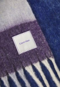 Deždana deka od vune u gradijentima ljubičaste, plave i sive boje s bijelom oznakom na kojoj je ispisano "Calvin Klein" ljubičastim koncem. Rubovi sa rese.