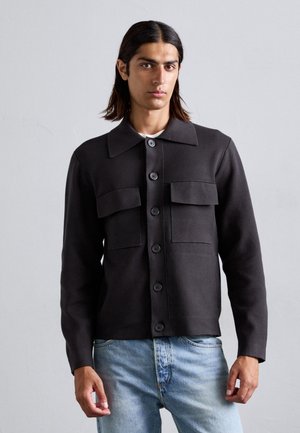 Svart knappjakke med krage, to brystlommer og strukturell stoff. Kombinert med lyseblå denimjeans. Enkelt design.