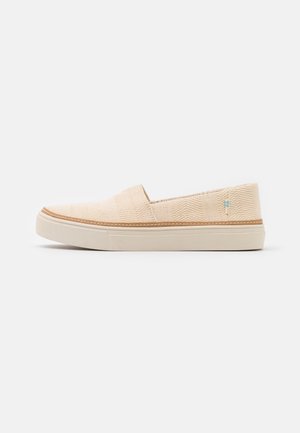 Beżowy sneaker wsuwany z teksturowanym materiałowym wierzchem, białą gumową podeszwą i małą niebiesko-białą metką po stronie pięty.