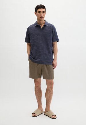 Man staat en draagt een getextureerd marineblauw poloshirt met korte mouwen, bruine shorts en beige instapsandalen tegen een effen witte achtergrond.