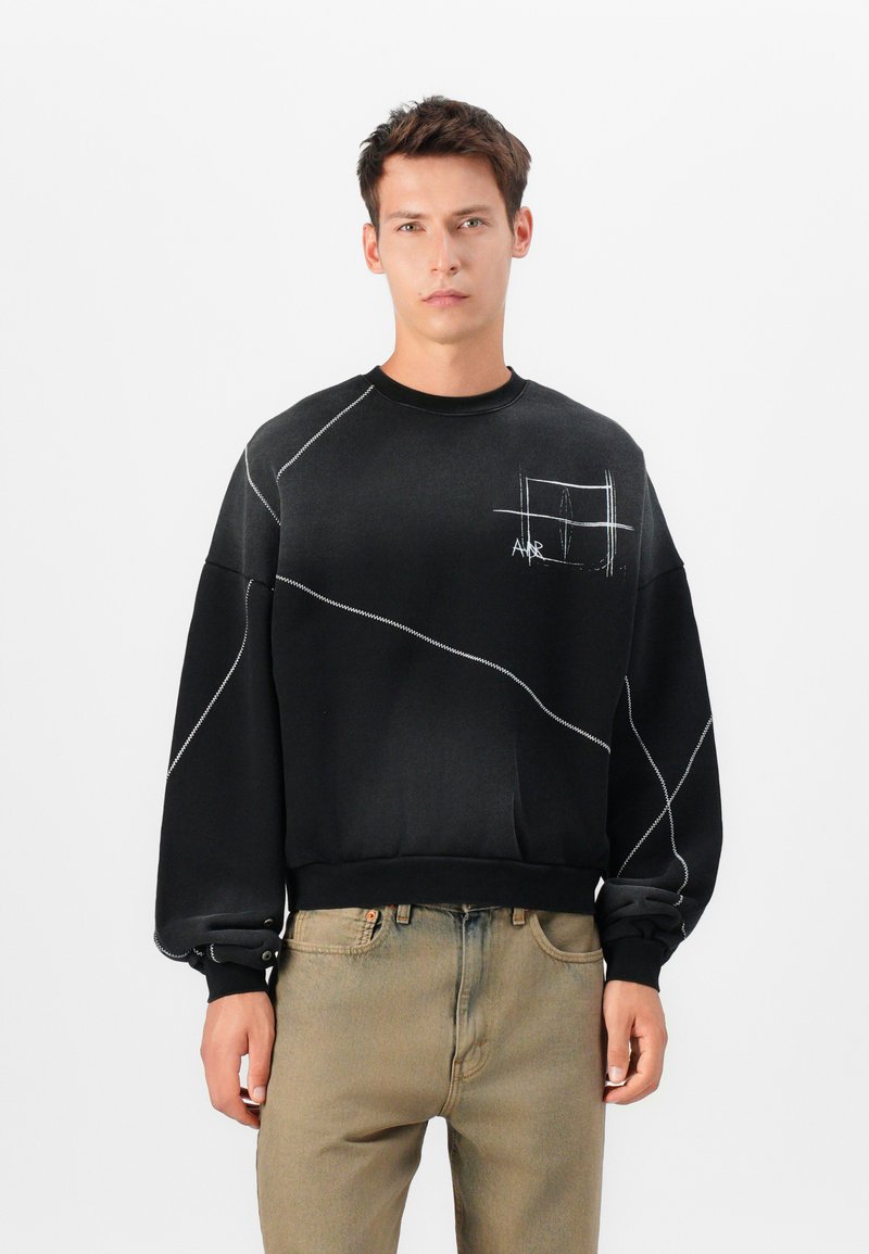 Sort sweatshirt med skrå hvid syning, rund halsudskæring og en brystlomme foran. Materialet ser blødt og struktureret ud.