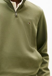Homme portant un pull-over olive vert à fermeture éclair quart avec un petit logo rouge, blanc et noir sur la poitrine gauche, chemise blanche en dessous.