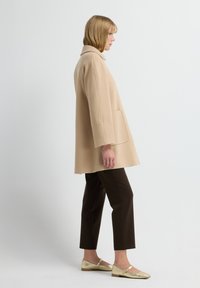 Beige oversized jas met een kraag en zakken, getextureerde mouwen, gecombineerd met bruine broeken en gouden platte schoenen. Eenvoudig en modern ontwerp.