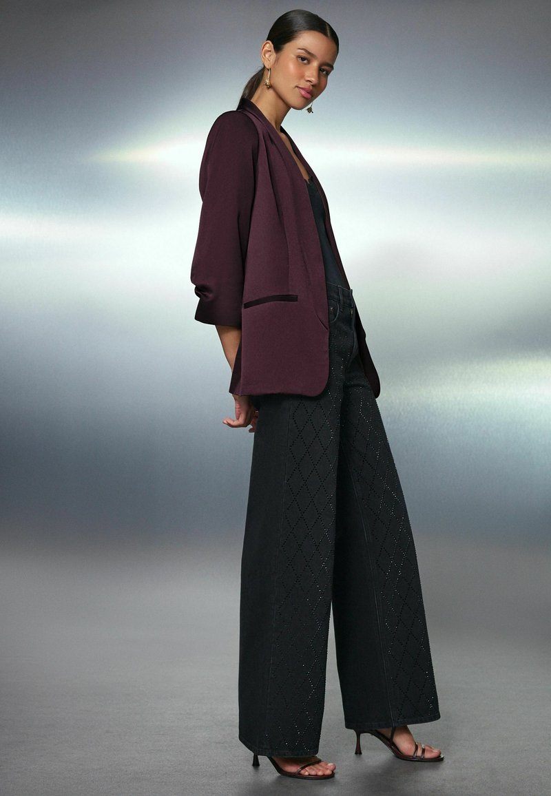 Donna che posa indossando pantaloni neri a gamba larga con ritagli a diamante, giacca bordeaux, top nero e sandali con tacco su sfondo grigio sfumato.