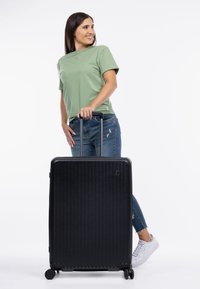 Valigia rigida nera con righe verticali, manico retrattile e 4 ruote. È in posizione verticale accanto a una persona con una maglietta verde.