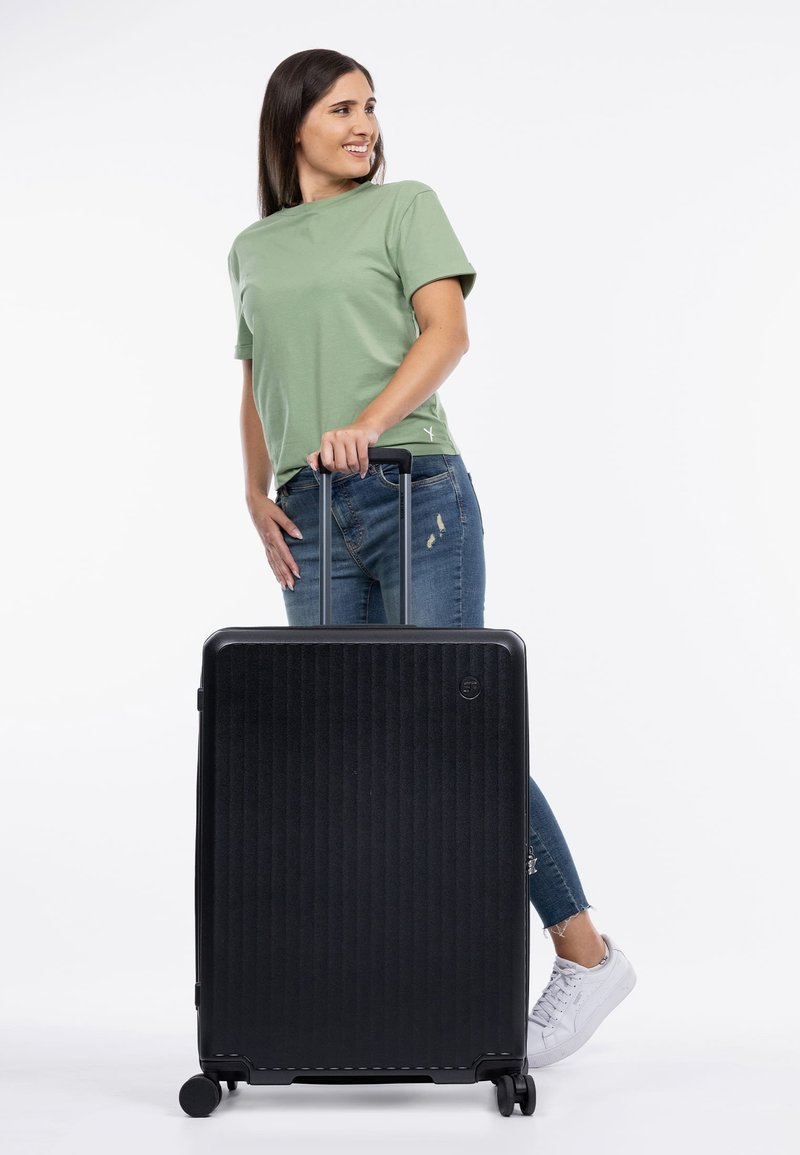Valigia rigida nera con righe verticali, manico retrattile e 4 ruote. È in posizione verticale accanto a una persona con una maglietta verde.