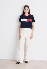 Marineblå bomulds T-shirt med rød og hvid stribet logo, parret med højtaljede cremefarvede jeans. Sorte spidse sko med dekorative detaljer.