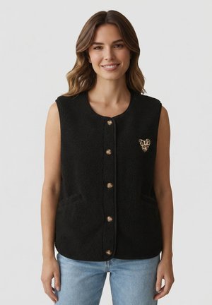 Donna sorridente, che indossa un gilet di pile nero senza maniche con bottoni e dettaglio a cuore con stampa leopardata, e jeans azzurri, su sfondo semplice.