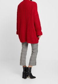 Suéter rojo de punto oversize con un patrón texturizado, combinado con pantalones cropped de cuadros grises y botas de tacón negro brillante hasta el tobillo.