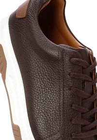 Zapatilla de cuero marrón con superficie texturizada, acentos en un marrón claro contrastante, suela de goma blanca y cordones marrones.