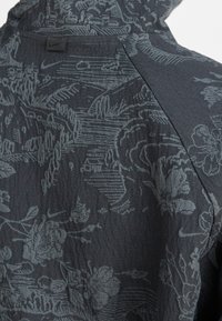 Veste à capuche Nike en tissu gris foncé avec un motif floral subtil, une surface texturée et un petit logo au niveau de l'encolure arrière.
