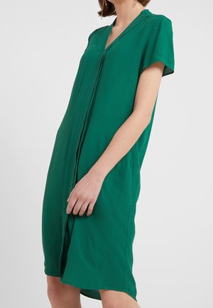 Kobieta w zielonej sukience z krótkim rękawem do kolan, z pionowymi plisami z przodu, stojąca na jednolitym tle.