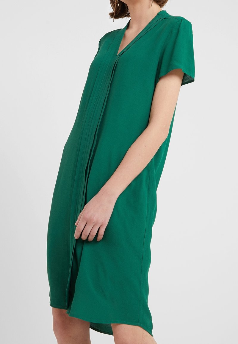Femme portant une robe verte à manches courtes, longueur genou, avec des plis verticaux sur le devant, debout devant un fond uni.