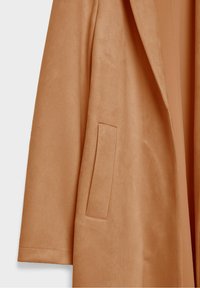 Manteau en daim couleur fauve avec un col à revers et une poche latérale unique. Le tissu présente une texture douce avec une finition lisse.