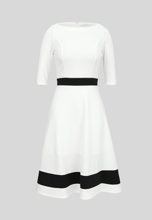 Hvit kjole til knelengde med trekvart lange ermer, svart midjebånd og svart horisontal stripe nær kanten på grå bakgrunn.