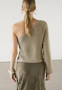 Pull en tricot beige avec un design asymétrique, comprenant une manche longue et une épaule dénudée. Porté avec une jupe texturée vert olive.