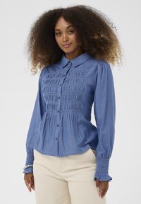 Vaaleansininen napillinen paita smockatut etuosat ja hihansuut, jossa leveä kaulus ja pitkät hihat, valmistettu pehmeästä, teksturoidusta kankaasta.