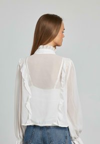 Weiße Chiffonbluse mit Rüschendetails, hohem Kragen und langen Ärmeln; kombiniert mit blauen Jeans. Das durchsichtige Material lässt die darunterliegende Schicht durchscheinen.