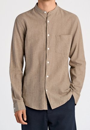 Homme portant une chemise beige à manches longues avec col montant et poche poitrine, associée à un pantalon foncé, debout les mains détendues.