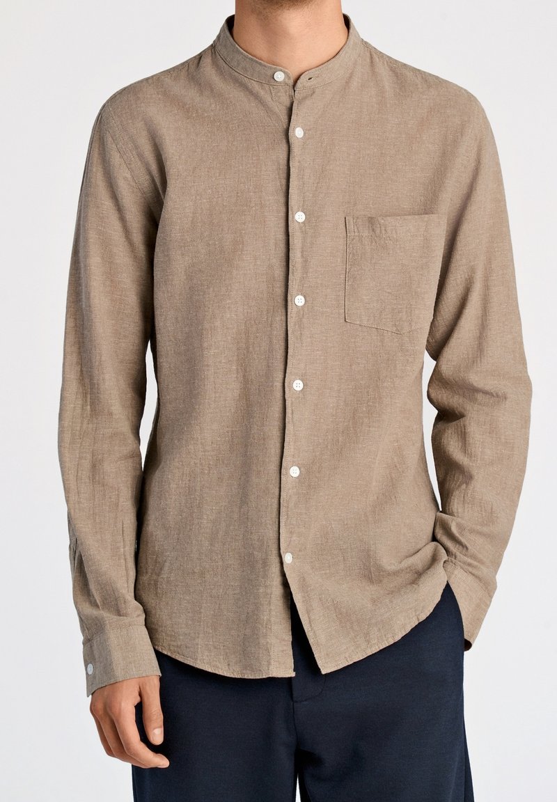 Homme portant une chemise beige à manches longues avec col montant et poche poitrine, associée à un pantalon foncé, debout les mains détendues.