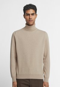 Beige polokragejumper tillverkad av mjuk stickad tyg. Har långa ärmar och ribbad nederkant. Slät textur med minimal dekoration.