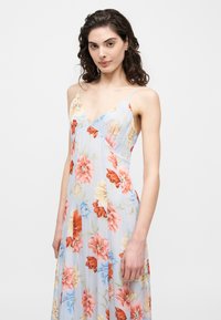 Νεαρή γυναίκα με σγουρά σκουρόχρωμα μαλλιά, που φοράει ένα ανοιχτό μπλε φλοράλ αμάνικο φόρεμα με λεπτές τιράντες, μπροστά σε απλό λευκό φόντο.