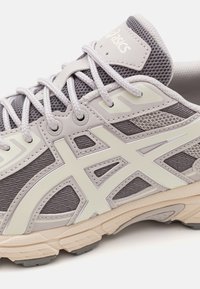 Scarpa sportiva grigia con tomaia in rete, caratterizzata da accenti bianchi e una suola con motivo. I lacci e il logo sono visibili. Dettagli texturizzati arricchiscono il design.