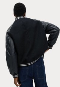 Chaqueta bomber negra con un cuerpo de forro polar texturizado, mangas de cuero brillante y puños y dobladillo de canalé a rayas blancas.