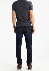 Dunkelblaue Jeans aus Denim mit einem verblassten Finish, schmaler Schnitt und Stickereien. Kombiniert mit einem grauen Kurzarmhemd und braunen Schuhen.