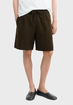 Muškarac odjeven u tamno smeđe teksturirane hlače s vezicama, bijelu košulju i crne cipele bez vezica, stoji s rukama u džepovima.