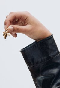 Ohrringe in Herz- und Pfeilform in goldfarbener Ausführung, zwischen Fingern gehalten, vor dem Hintergrund eines schwarzen Lederjackenärmel.