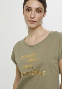 Olivgrünes T-Shirt mit kurzen Ärmeln aus weichem Stoff, das in lässiger Schriftart mit goldfarbener Folienbeschriftung "create your own sunshine" bedruckt ist.