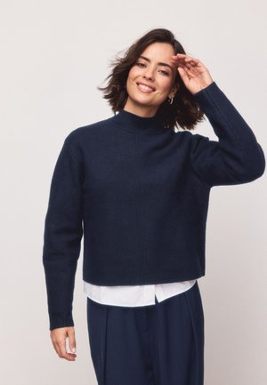 Kvinde med kort mørkt hår, der smiler, iført en mørkeblå sweater over en hvid skjorte og matchende mørkeblå bukser, poserer med en hånd nær hovedet.
