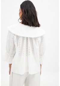 Blusa bianca con un ampio colletto rotondo, motivo ad occhiello ricamato, maniche a sbuffo e retro arricciato. Tessuto leggero con un design fluente.