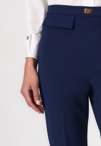 Σαλοπέτες σε σκούρο μπλε χρώμα, φτιαγμένες από μαλακό ύφασμα, με επίπεδο μπροστινό μέρος και χρυσό κούμπωμα. Λεπτομέρεια σε τσέπη.