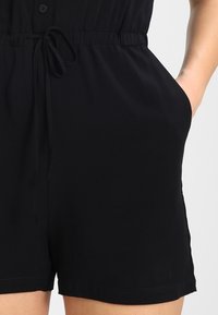 Combinaison noire avec un devant boutonné, une taille élastique avec cordon de serrage et des poches latérales. Tissu lisse avec une coupe décontractée et des jambes courtes.