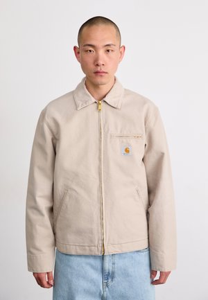 Veste en toile beige avec une fermeture éclair dorée, col montant et poches latérales ; possède une étiquette sur la poche poitrine. Portée par-dessus un jean bleu clair.