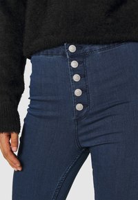 Primer plano de una persona que lleva vaqueros de tiro alto de color azul oscuro con cinco botones plateados visibles y una manga de suéter negro de felpa.