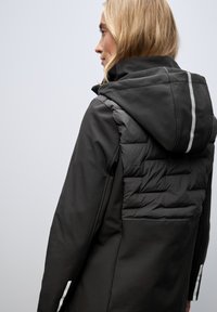 Black jacket featuring a puffer-style back, hood, and reflective strip details. Material appears smooth with a structured fit.