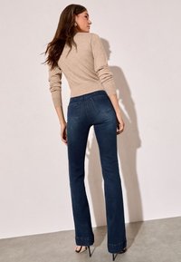 Smėlio spalvos aptemptas megztinis derinamas su tamsiai mėlynais išplatėjusiais džinsais. Džinsai turi kišenes nugaroje ir šiek tiek tekstūruotą audinį.